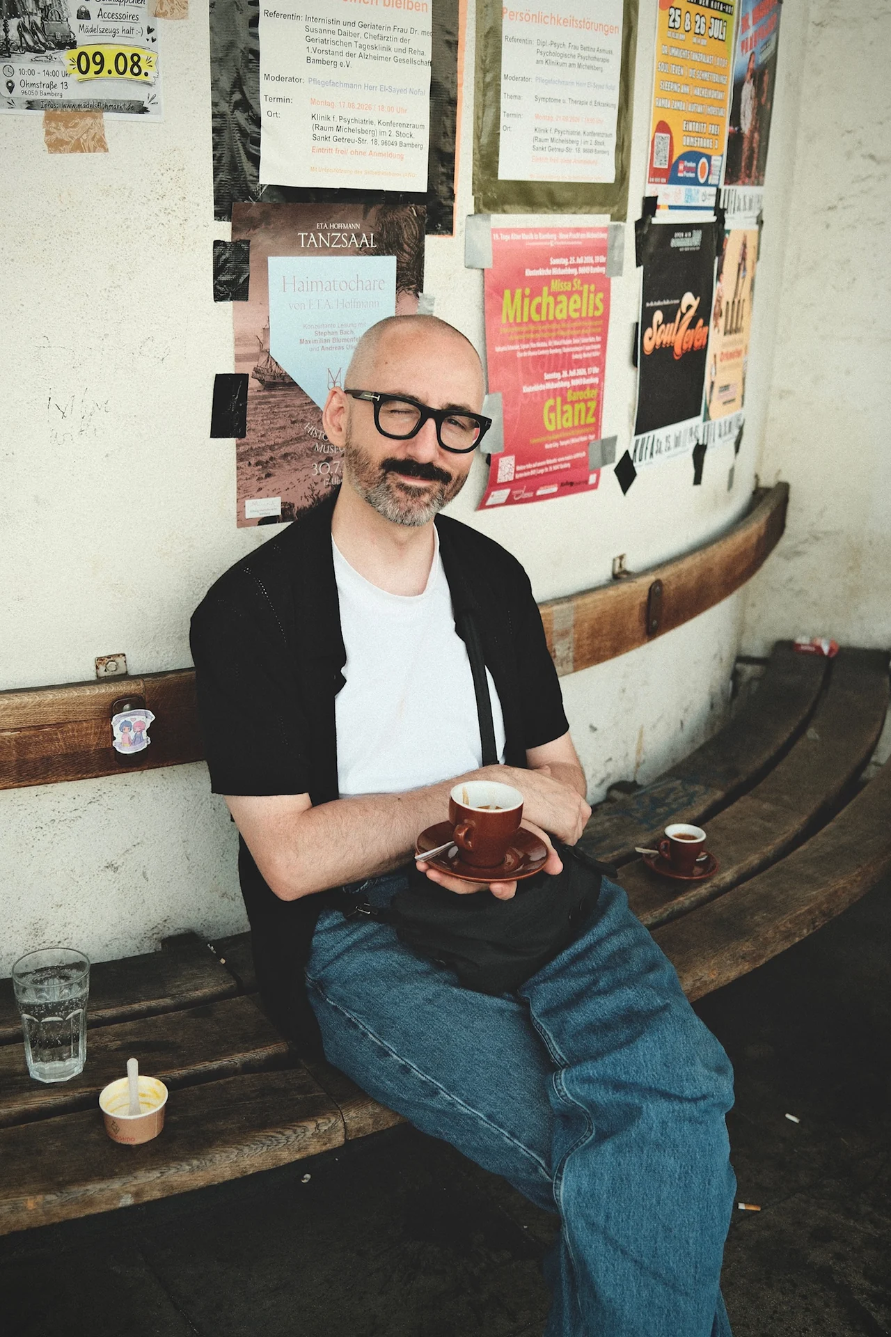 Ein Mann mit Brille und einer Kaffeetasse sitzt auf einer Holzbank vor Konzertplakaten.