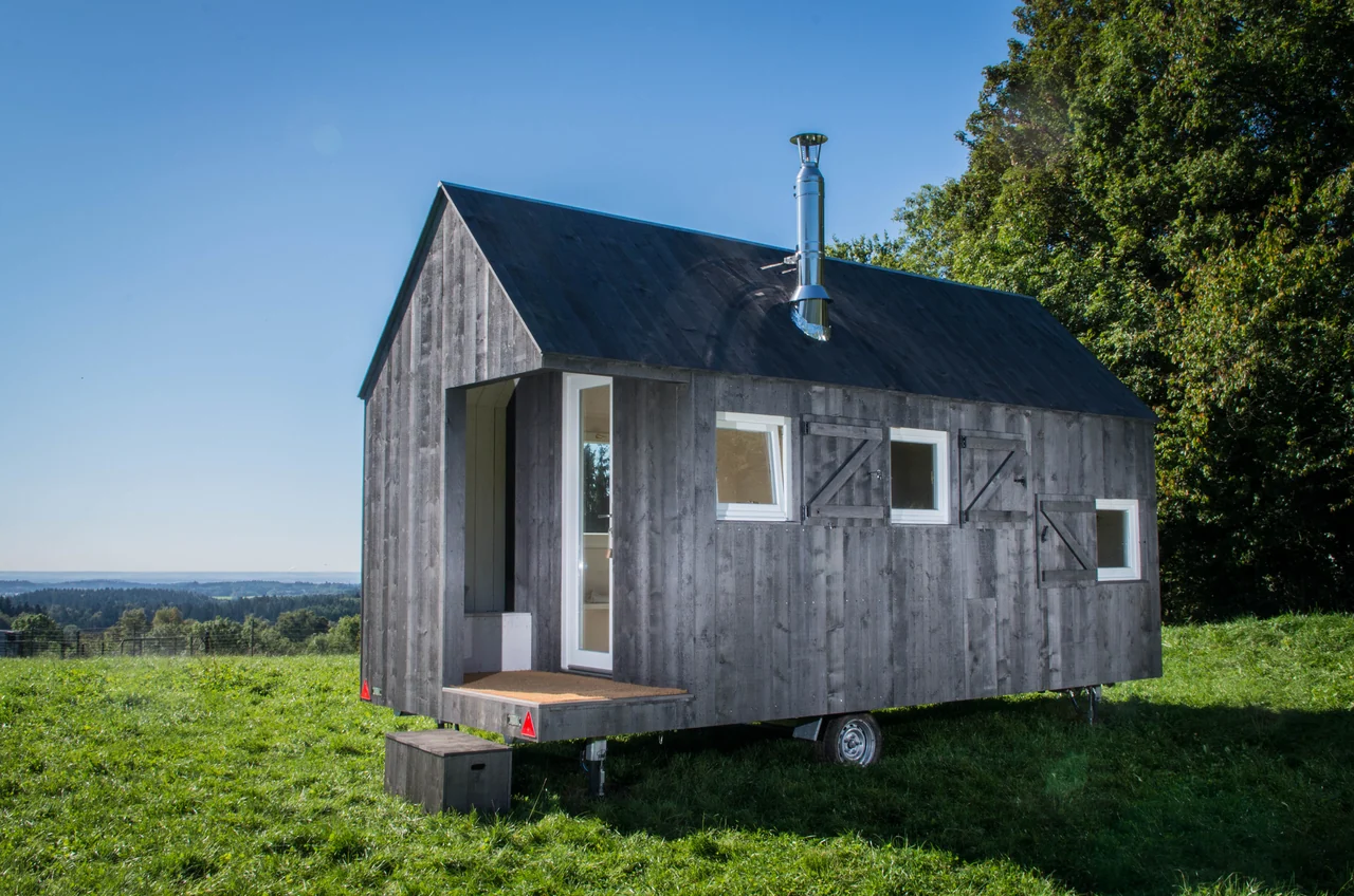 Das mobile Scheunenhäuschen HYT am Wildberghof Buchet in Dreiviertelansicht auf einer Wiese, mit Holzfassade, offenem Eingang, Ofenrohr und sichtbarem Fahrwerk.
