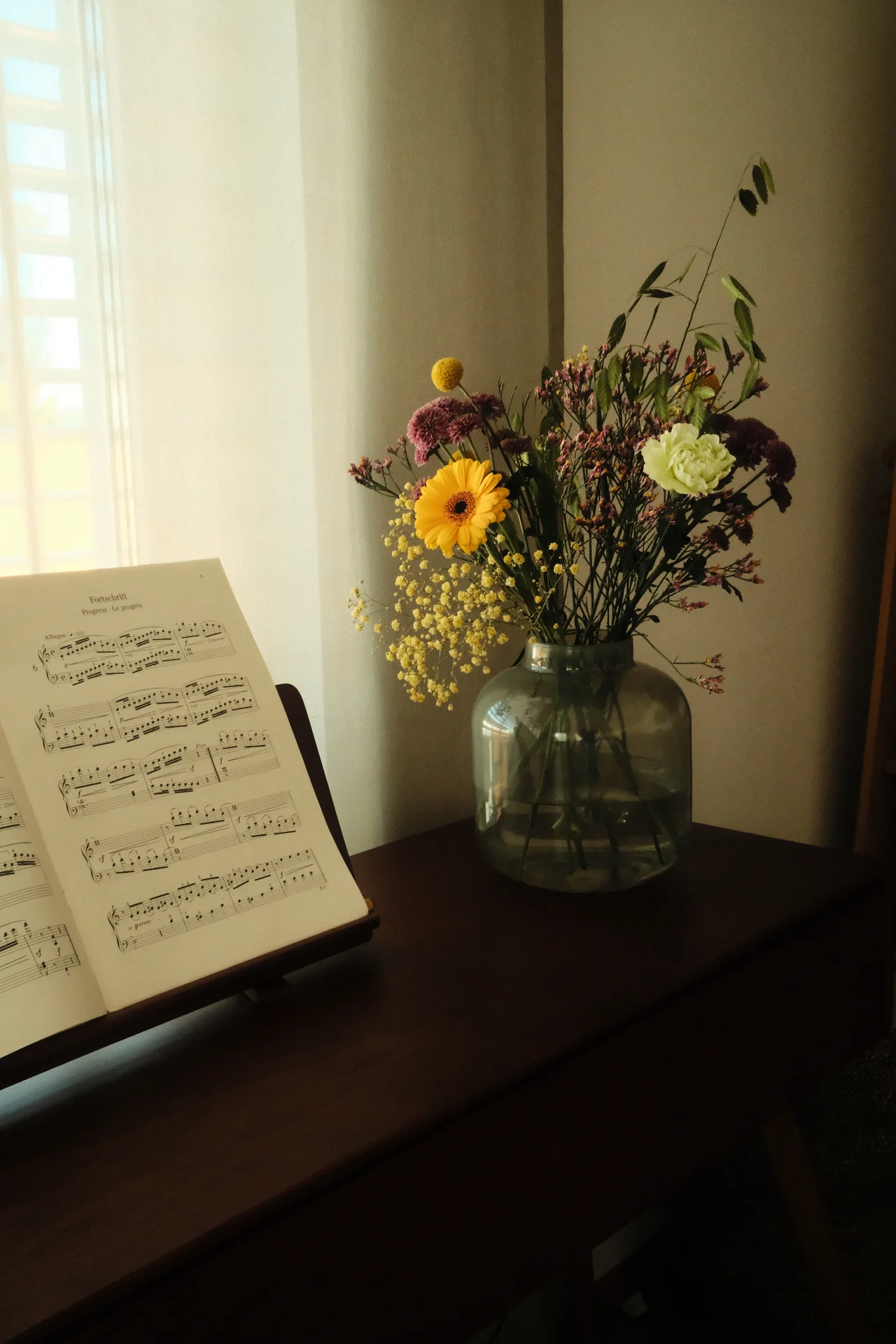 Noten auf einem Klavier neben einer Blumenvase im Fensterlicht.