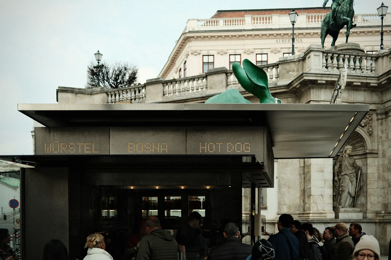 Ein Würstelstand mit Gästen vor einer historischen Fassade und einer grünen Hasenskulptur.